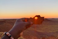 Hand holding rock with heart-shaped hole framing sunset over desert landscape